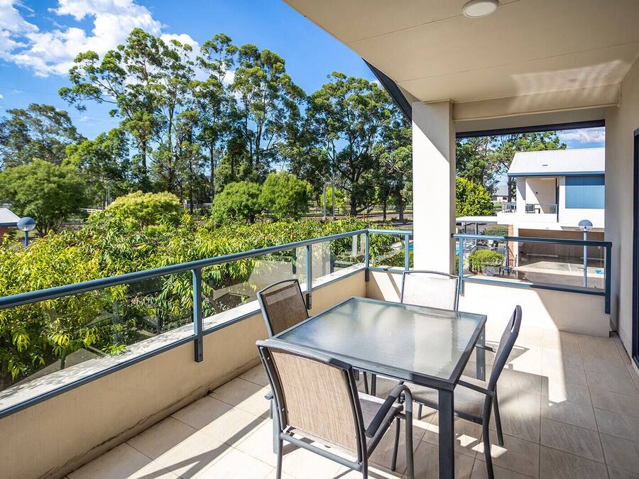 Un balcon avec une table et des chaises en verre, une rambarde en métal et une vue sur les arbres et les bâtiments.