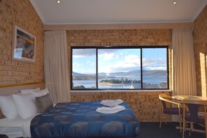 Habitación de hotel con un ventanal que ofrece vistas a un lago y montañas, una cama con cubrecama azul estampado, una pequeña mesa con sillas y un cuadro en la pared.