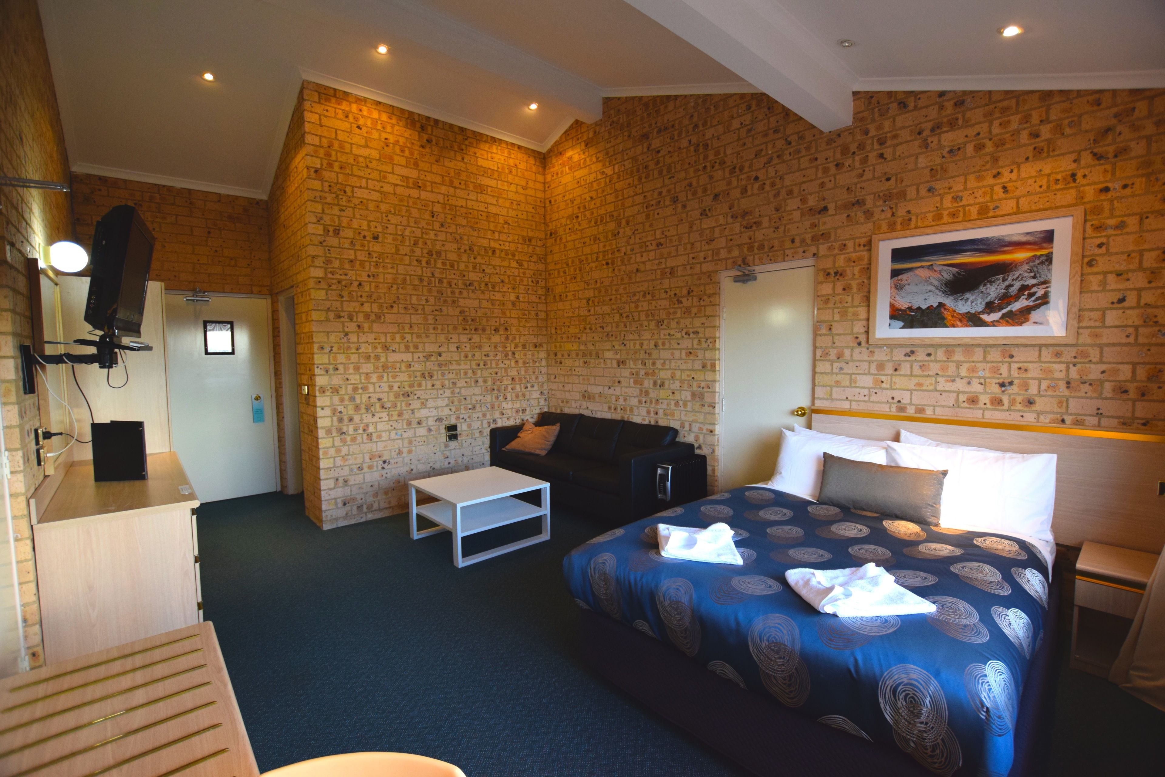 Habitación de hotel con pared de ladrillo, cama con colcha azul estampada, mesita de noche blanca, sofá negro y una pequeña mesa blanca.