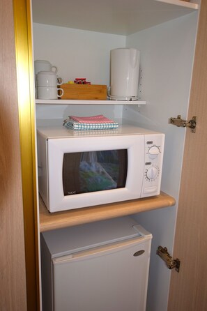 Una kitchenette compacta con microondas, un refrigerador pequeño y un estante con cafetera, hervidor de agua y una caja de pañuelos.