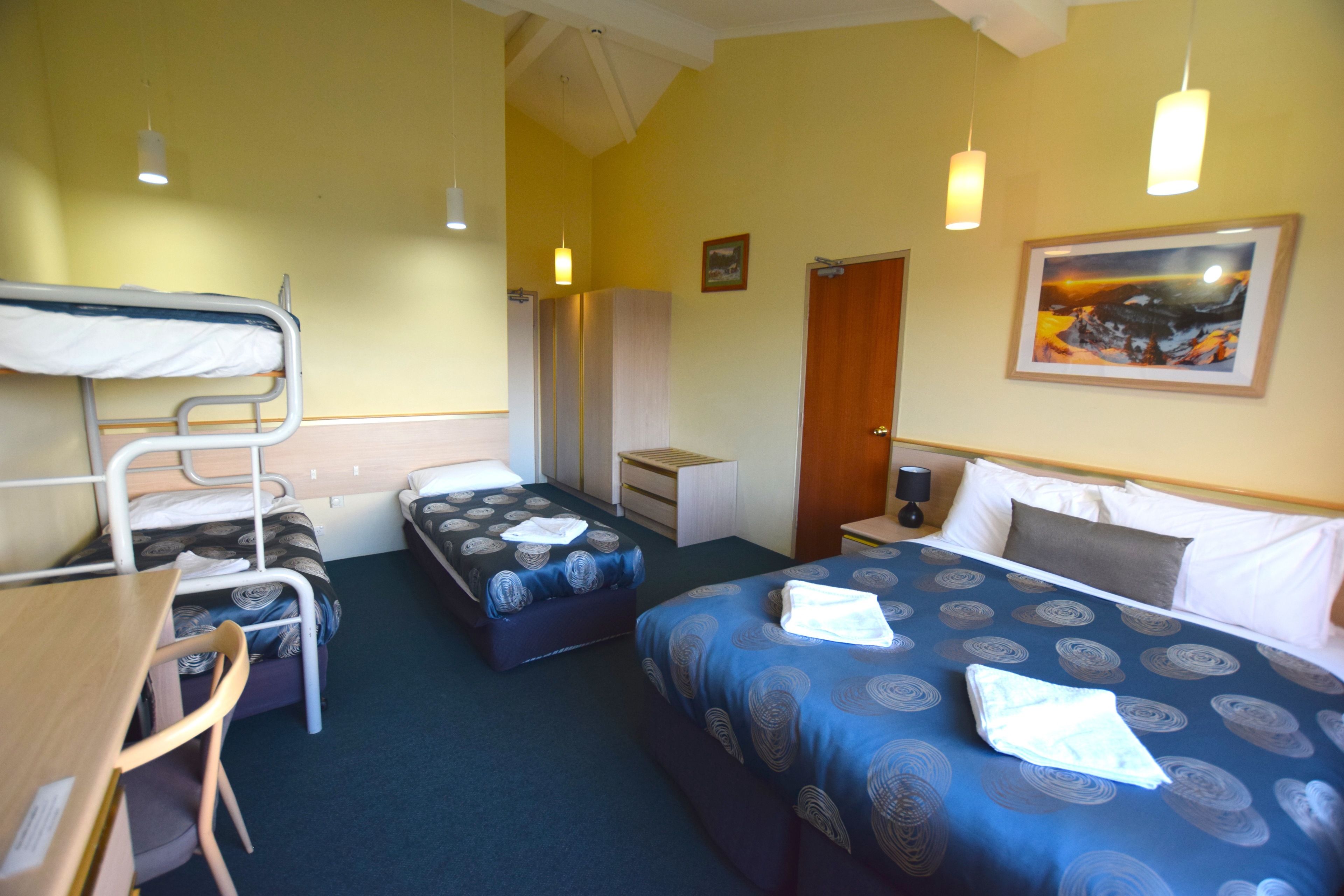 A hotel room with bunk beds, a desk, a chair, a wardrobe, a bedside table, and a bed with a patterned bedspread.