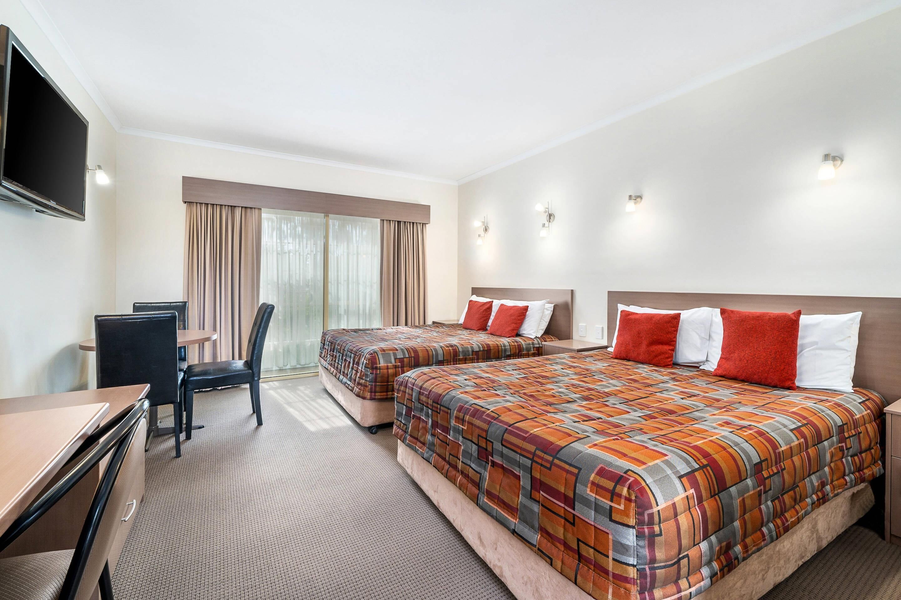 A hotel room with two beds, a desk, a chair, a TV, and a large window with curtains.