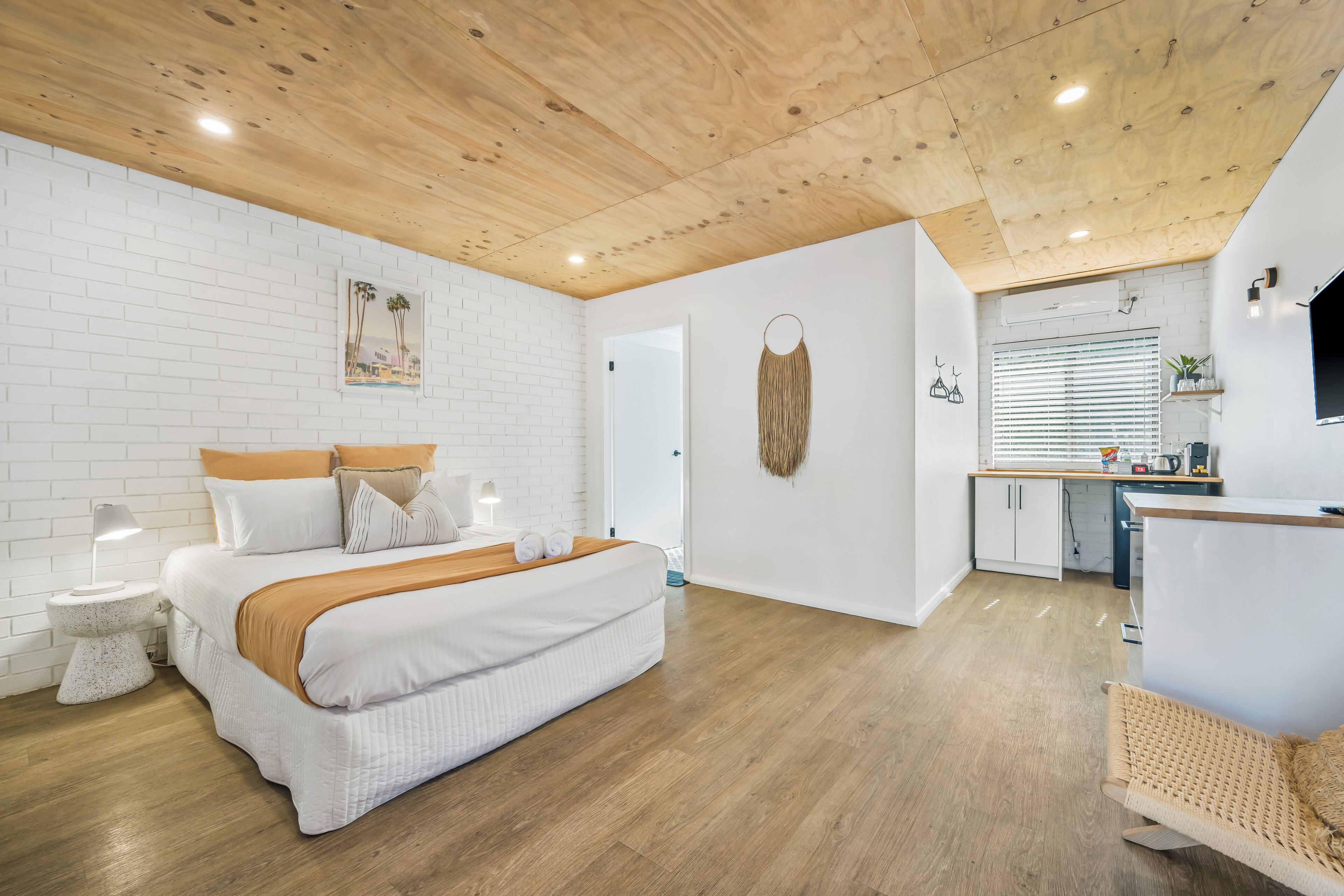 Une chambre moderne avec un grand lit, des tables de chevet, un plafond en bois et un coin cuisine équipé d’un comptoir et d’appareils électroménagers.