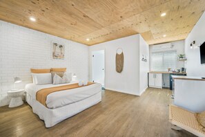 A modern bedroom with a large bed, bedside tables, a wooden ceiling, and a kitchen area with a counter and appliances.