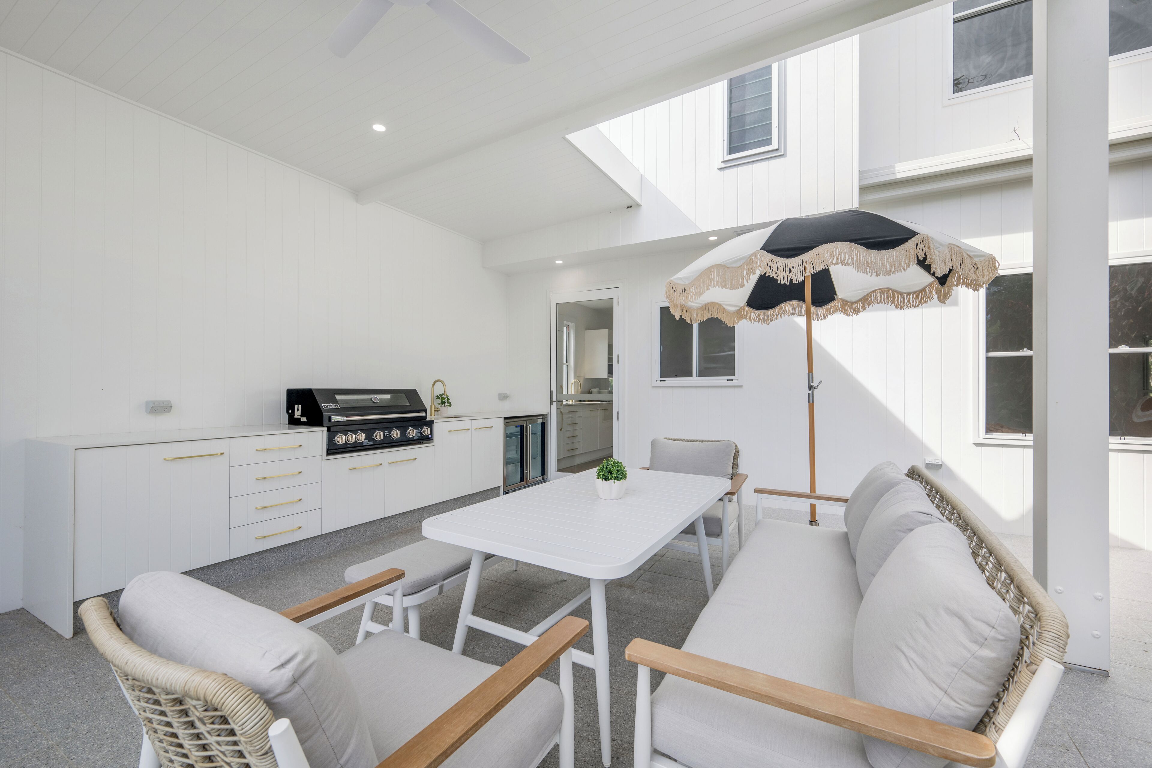 Een moderne keuken met witte kasten en een ingebouwde grill, een eethoek met een witte tafel en rieten stoelen, en een groot raam met een decoratieve parasol.