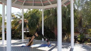 A covered outdoor area with a wooden deck, white pillars, and a person performing a yoga pose on a blue mat.