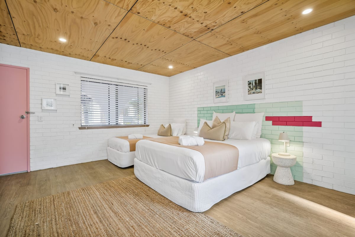 Quarto com teto de madeira, paredes de tijolo branco, porta rosa, janela com persianas, cama com roupa de cama branca, mesa de cabeceira com abajur e um tapete pequeno.