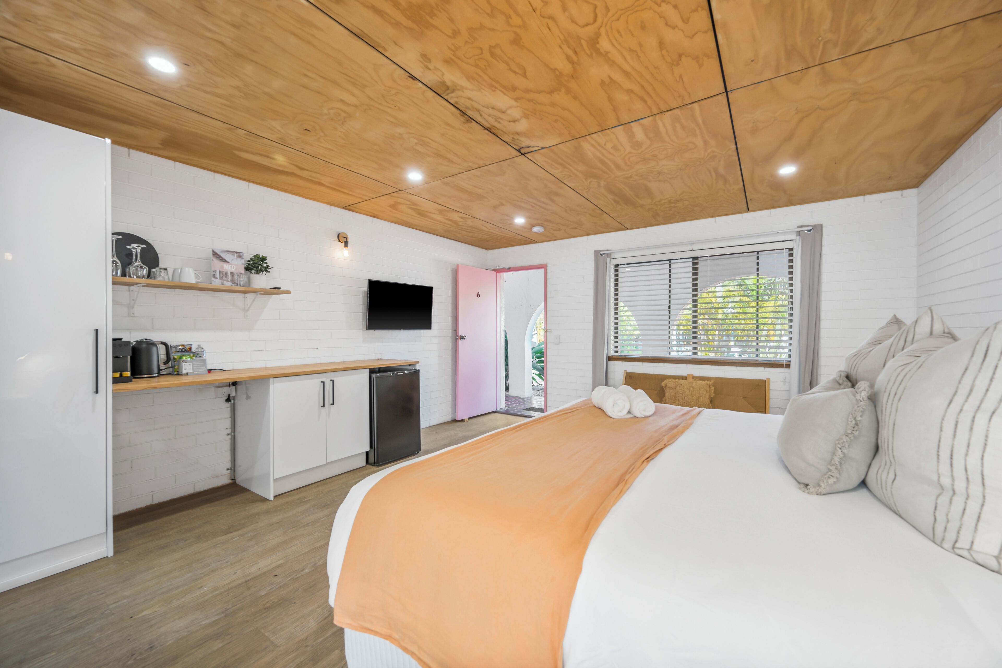 A modern bedroom with a wooden ceiling, a bed with an orange blanket, a TV, and a kitchenette with white cabinets and a pink door.