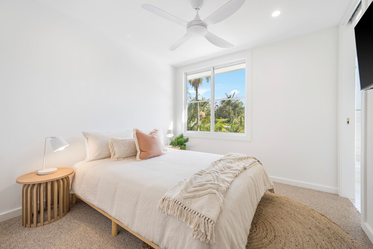 A bedroom with a bed, a bedside table, a lamp, a ceiling fan, a window with a view of greenery, and a TV mounted on the wall.