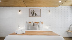 A modern hotel room with a wooden ceiling, white brick walls, a large bed with white bedding and striped pillows, and a framed cactus artwork above the bed.