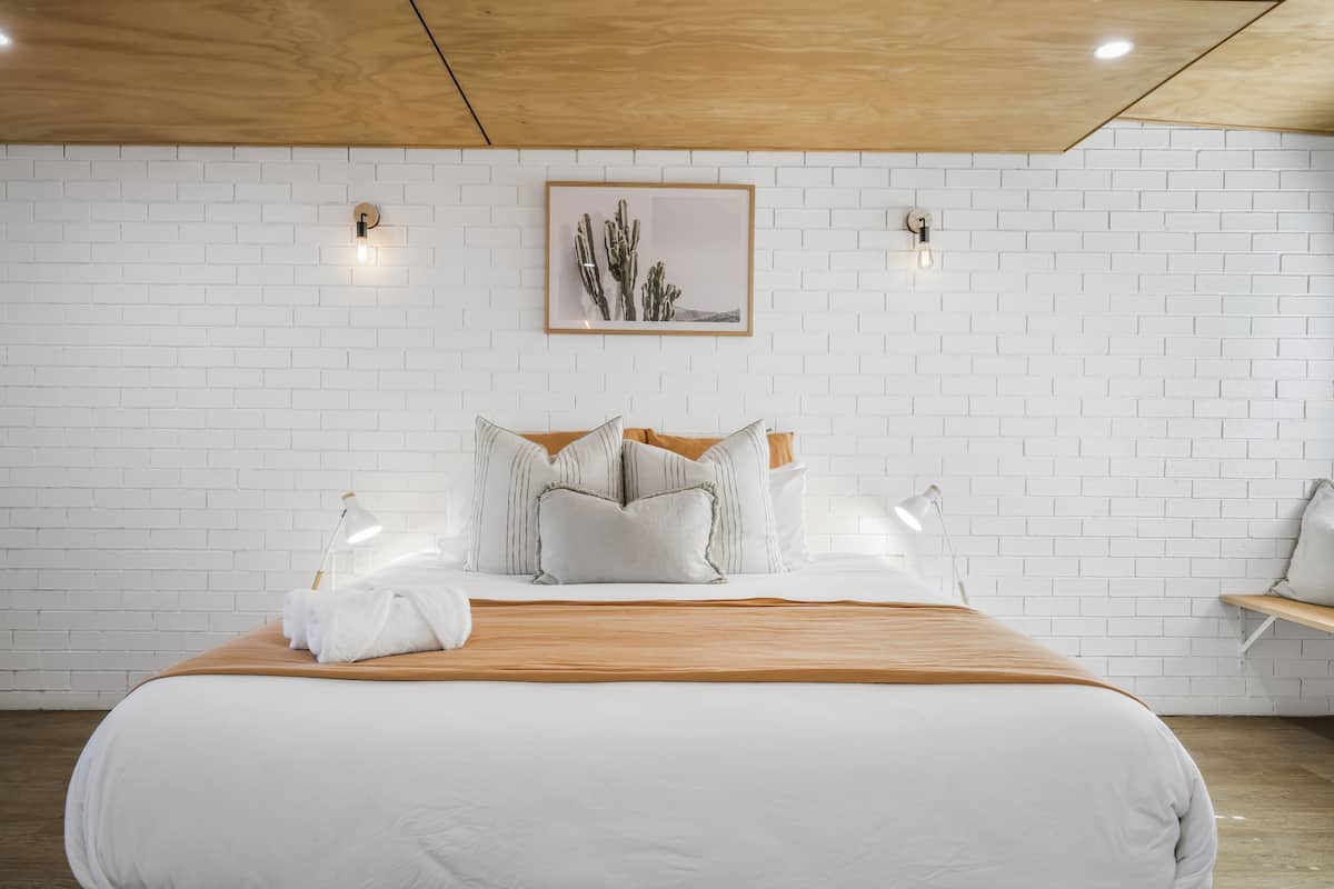 Quarto de hotel moderno com teto de madeira, paredes de tijolo branco, uma cama grande com roupa de cama branca e almofadas listradas, e uma ilustração de cactos emoldurada acima da cama.