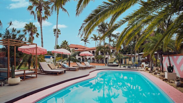 A pool area with lounge chairs and umbrellas, surrounded by palm trees and a building with a red roof.