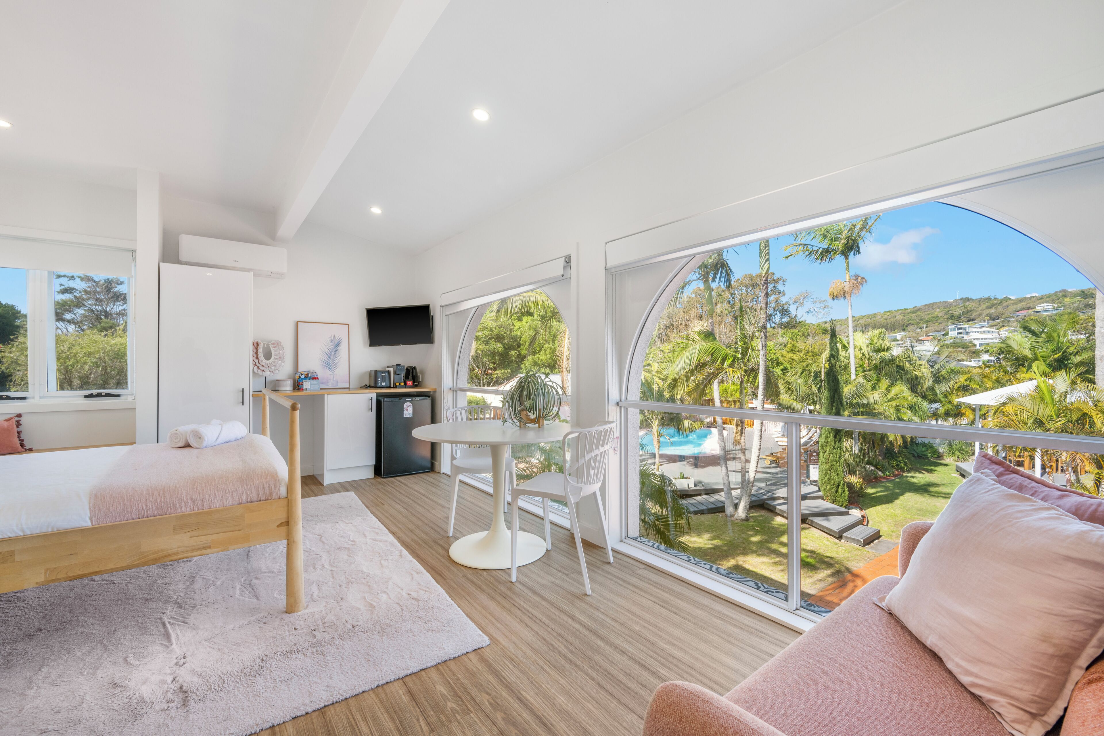 A modern bedroom with a large window, a bed, a desk, and a view of a pool and greenery.