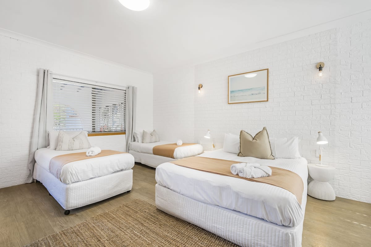 Quarto de hotel com duas camas, parede de tijolos brancos, um quadro emoldurado e uma pequena mesa de apoio com uma luminária.