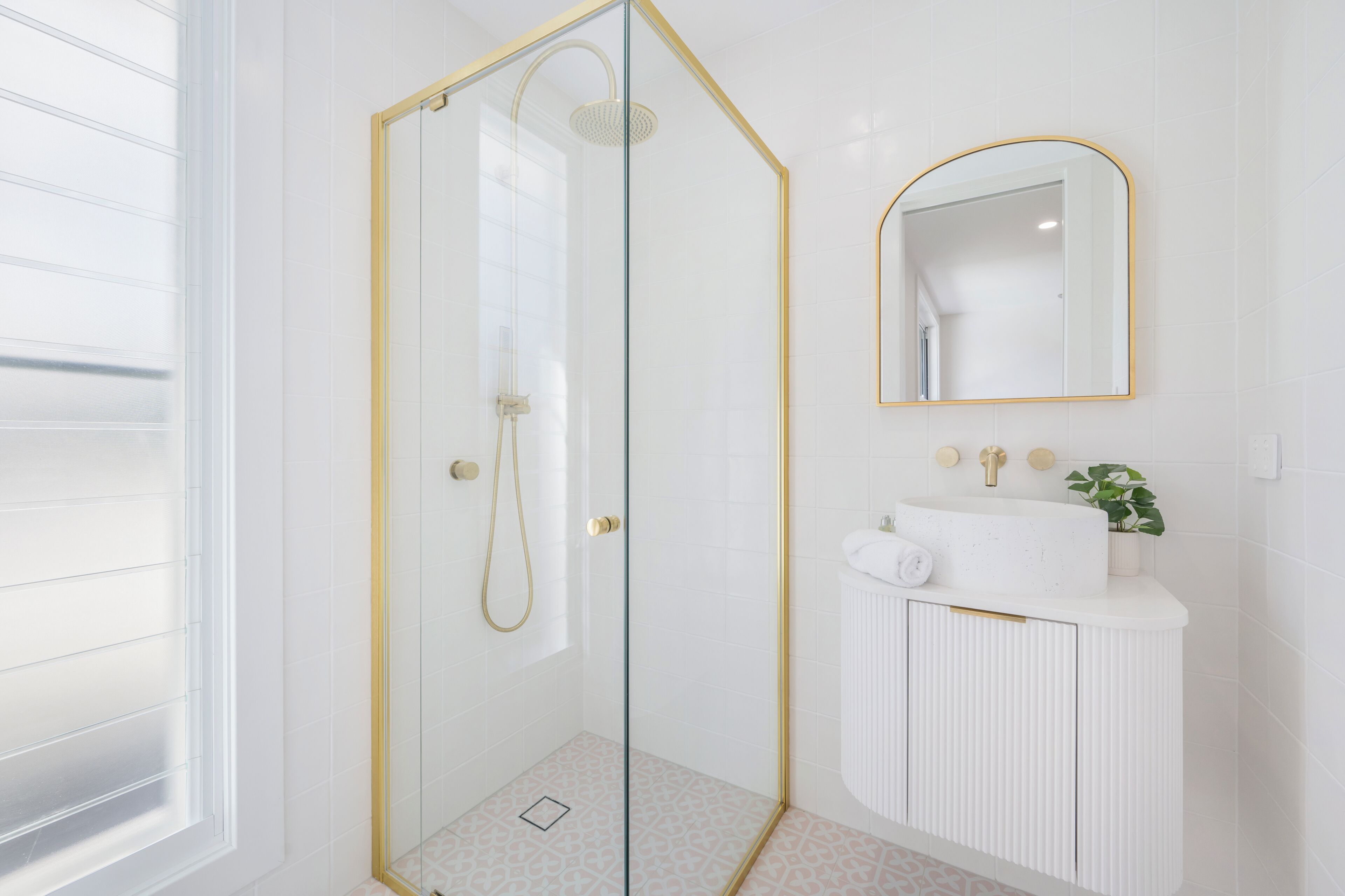 Un baño moderno con ducha envuelta en vidrio, tocador blanco con grifo dorado y espejo con marco dorado.