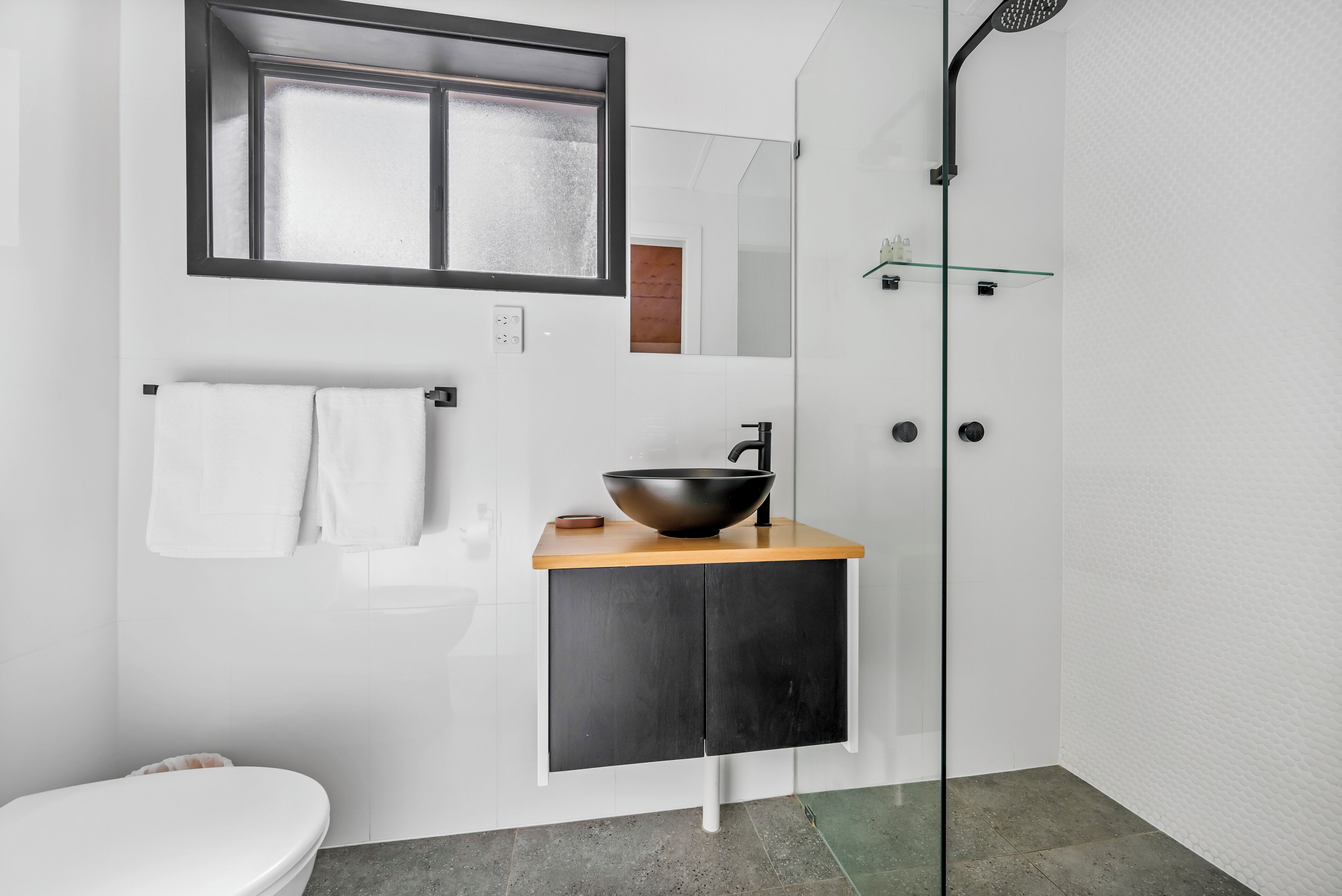 A modern bathroom with a large mirror, a black vessel sink, a white toilet, and a glass shower enclosure.