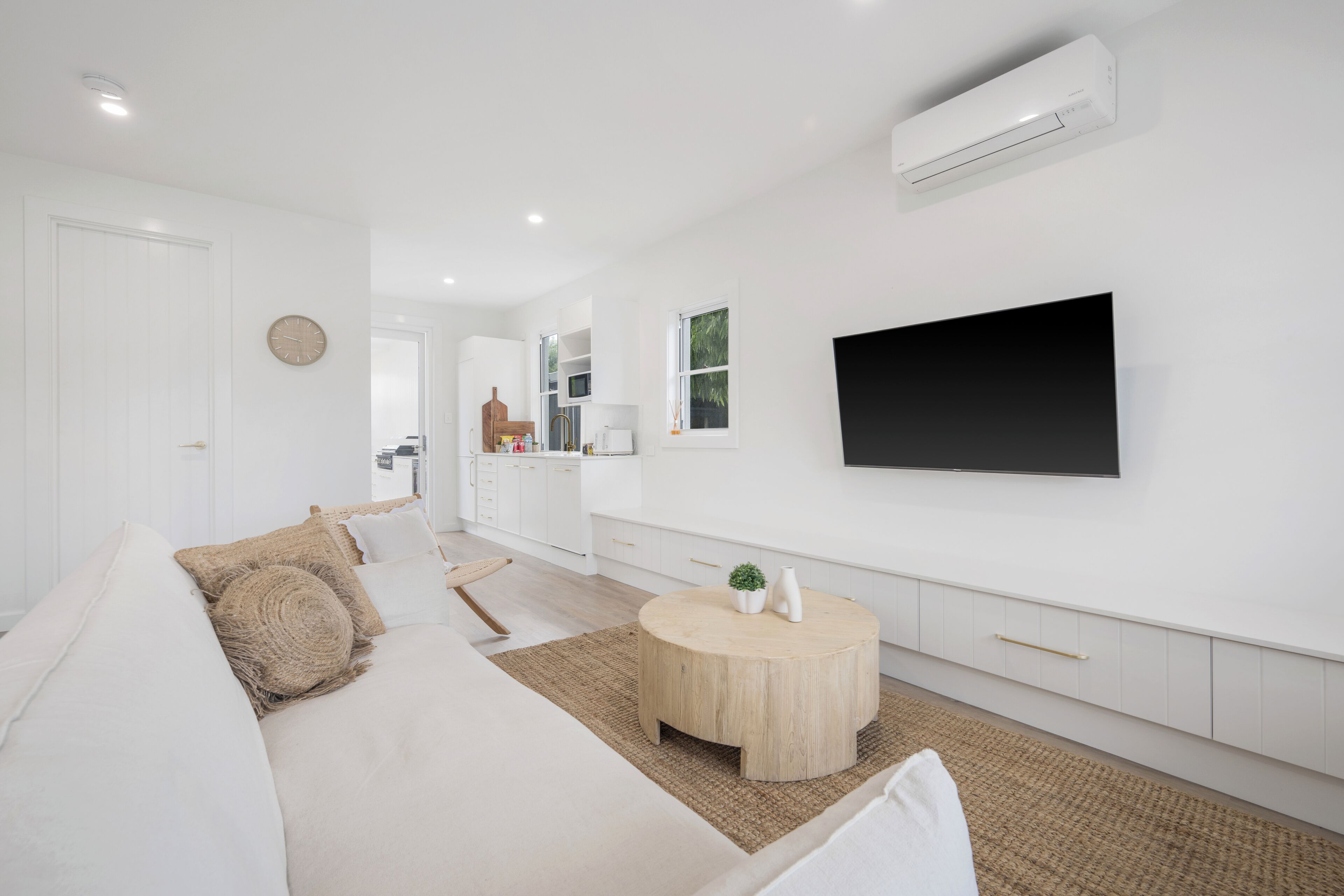 A modern living room with a sofa, a round coffee table, a flat-screen TV mounted on the wall, and a kitchen area with white cabinets and a window.