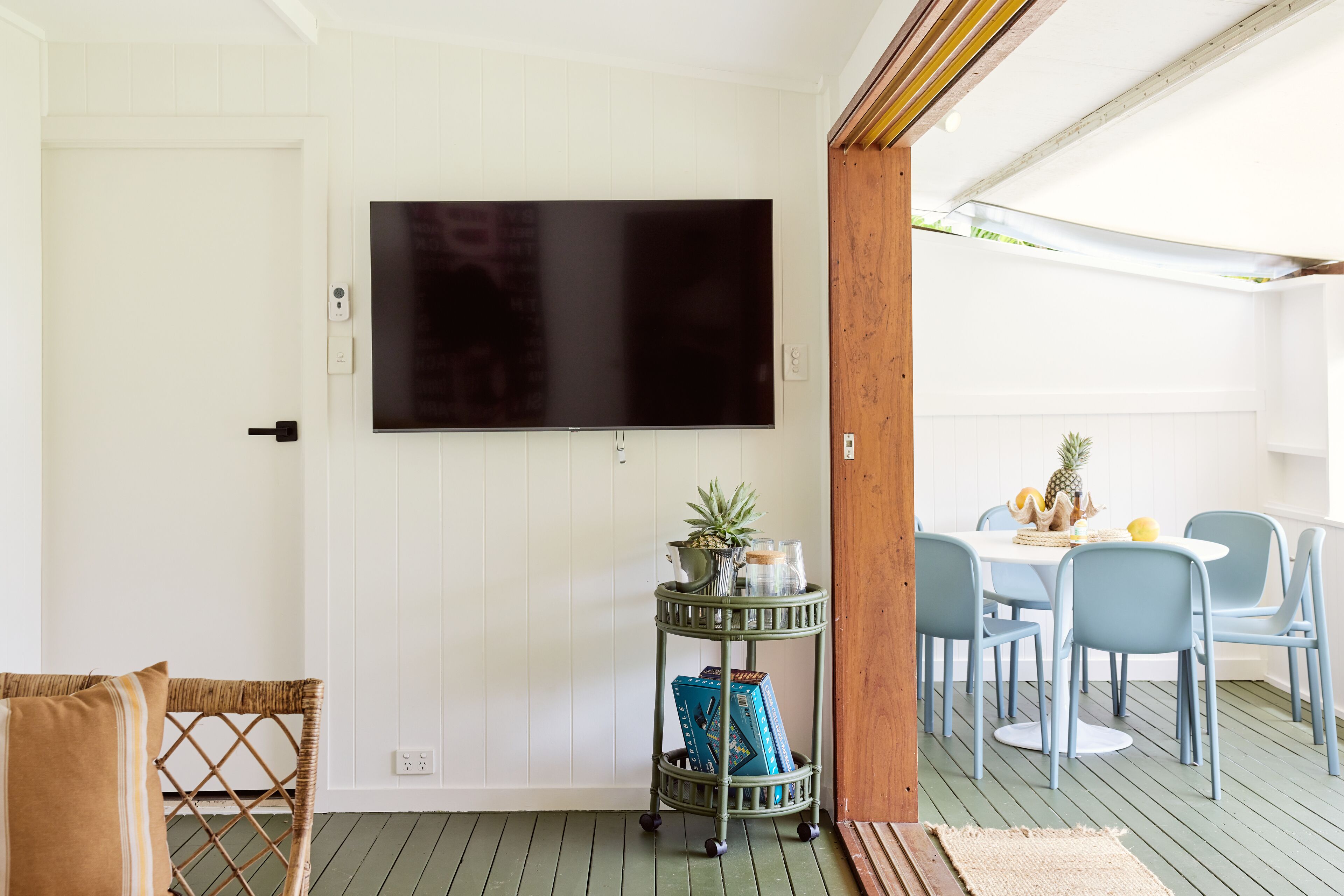 Una habitación con un televisor de pantalla plana montado, una silla de mimbre, un pequeño carrito metálico con libros y una planta, y un comedor con mesa y sillas.