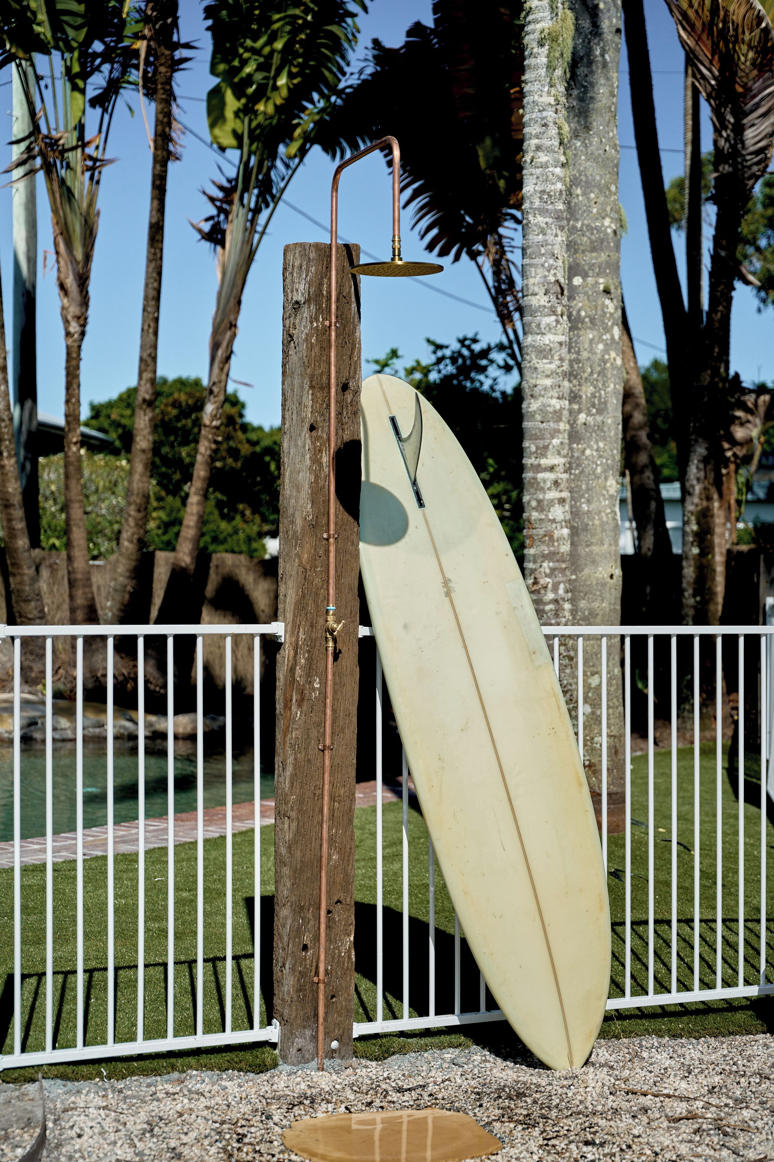 A surfboard leaning against a wooden post with a metal hook, surrounded by a white fence and palm trees.