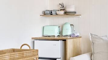 A kitchenette with a white refrigerator, a teal toaster, and a kettle on a wooden countertop. There are shelves above holding jars and bowls. A wicker basket and a beige sofa are also visible.