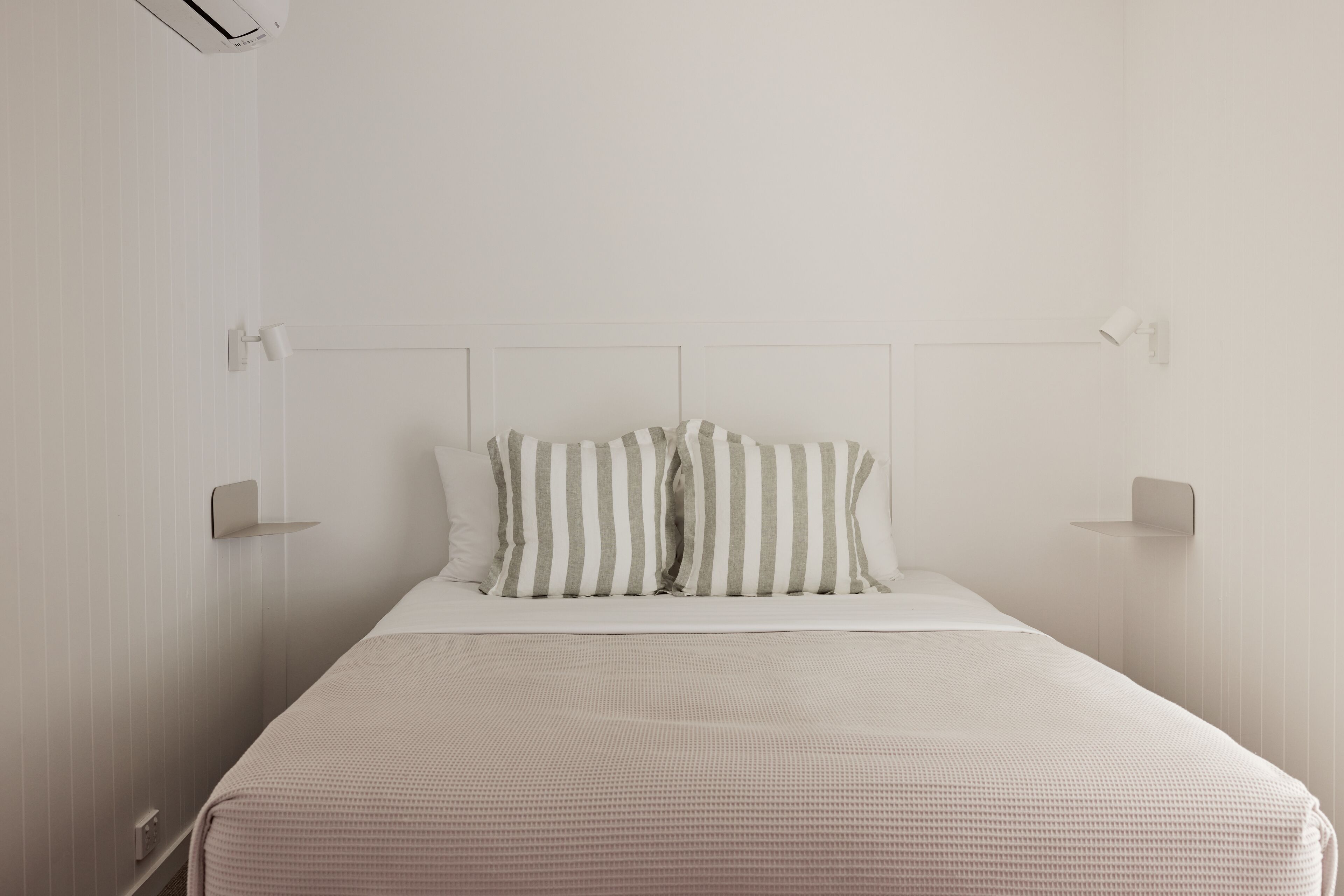 Une chambre à coucher au style minimaliste, avec un mur blanc uni, un lit simple agrémenté d’un coussin à rayures blanches et grises, et une tête de lit sobre.