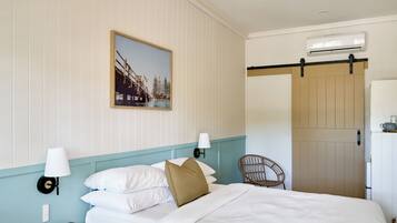 A hotel room with a bed, two wall-mounted lamps, a remote control, a picture on the wall, a wicker chair, and an air conditioning unit.