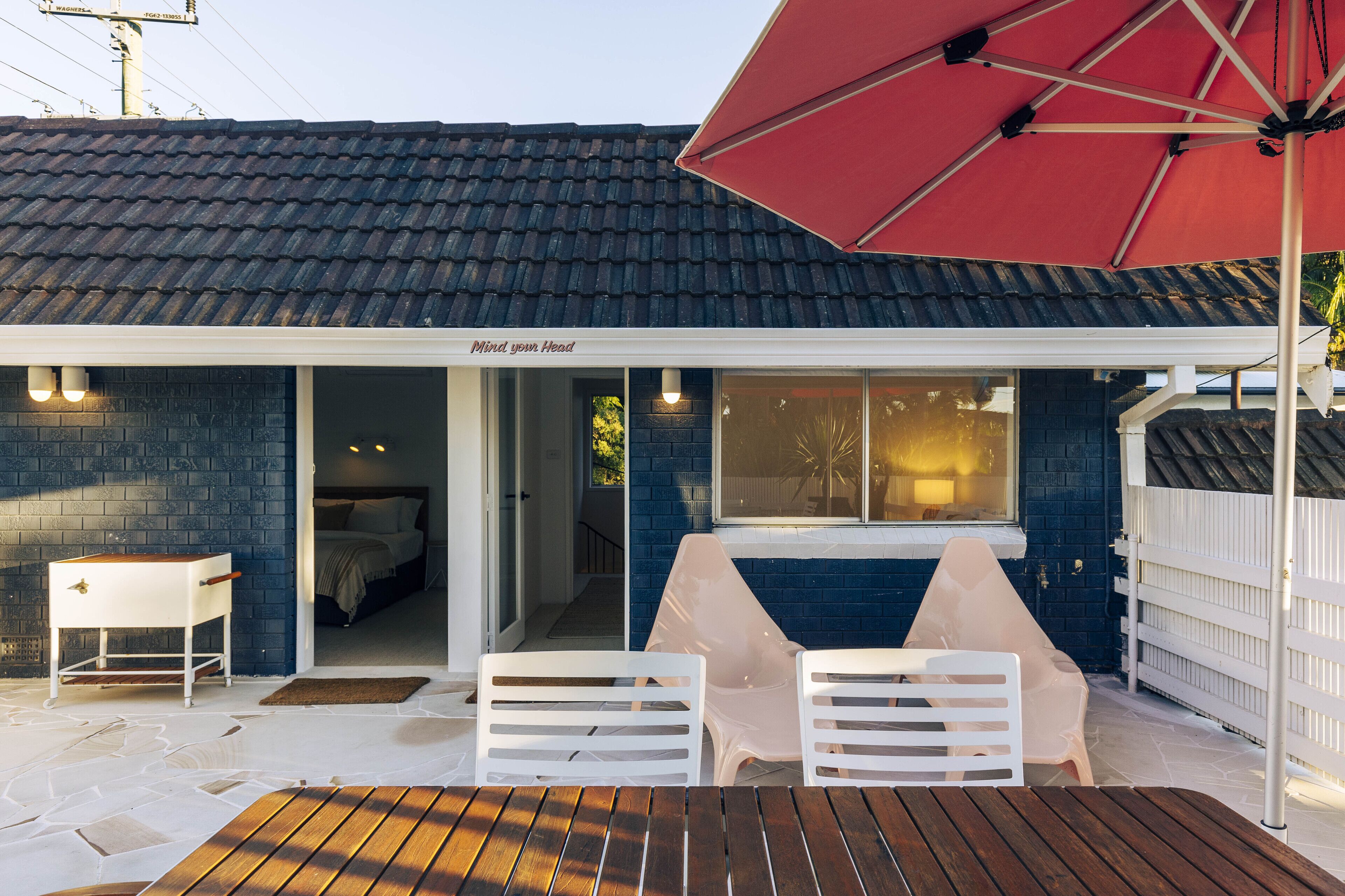 A patio area with a wooden table, two white chairs, a red umbrella, and a white fence.
