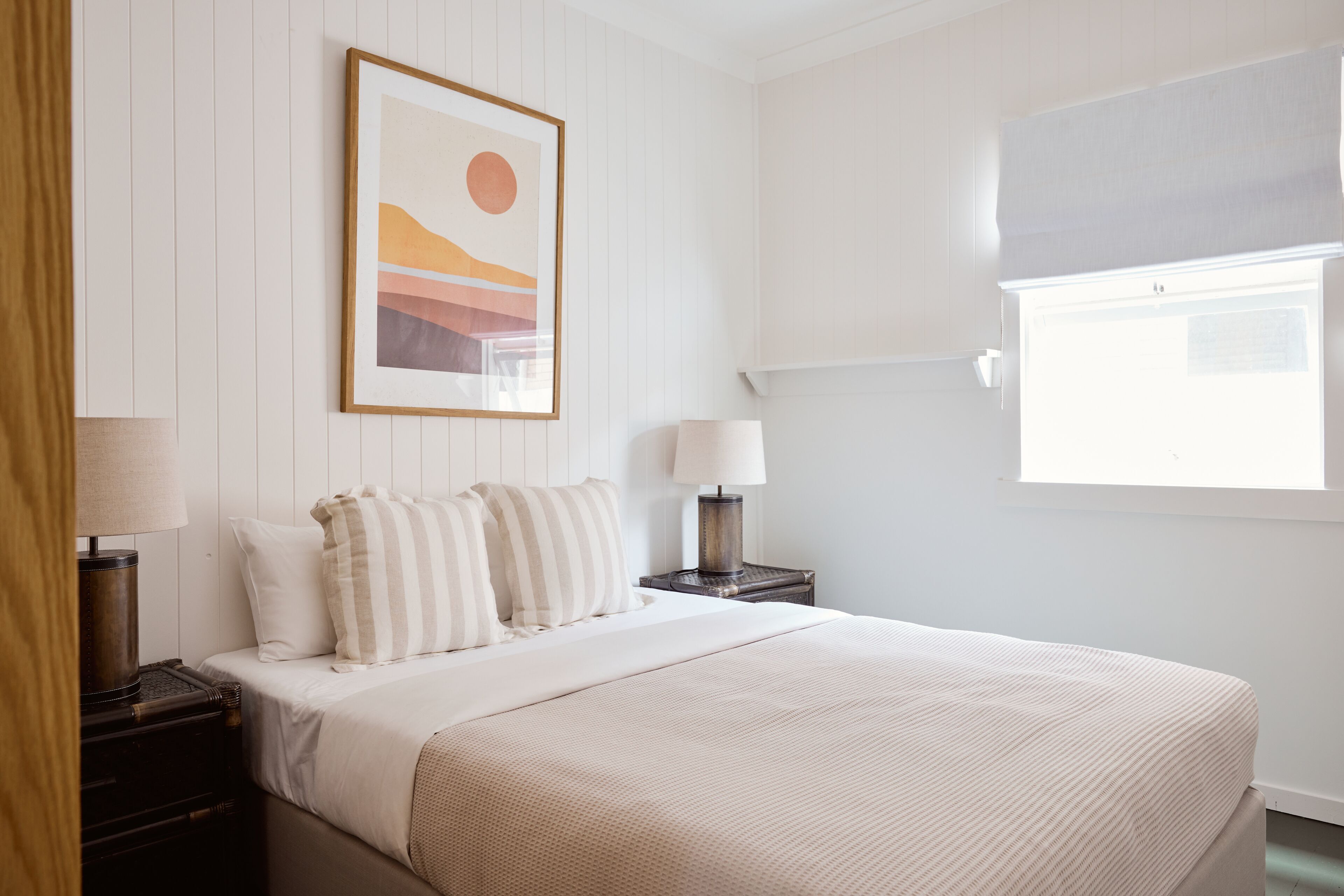 A neatly arranged bedroom with a bed, pillows, a framed picture, bedside lamps, and a window.