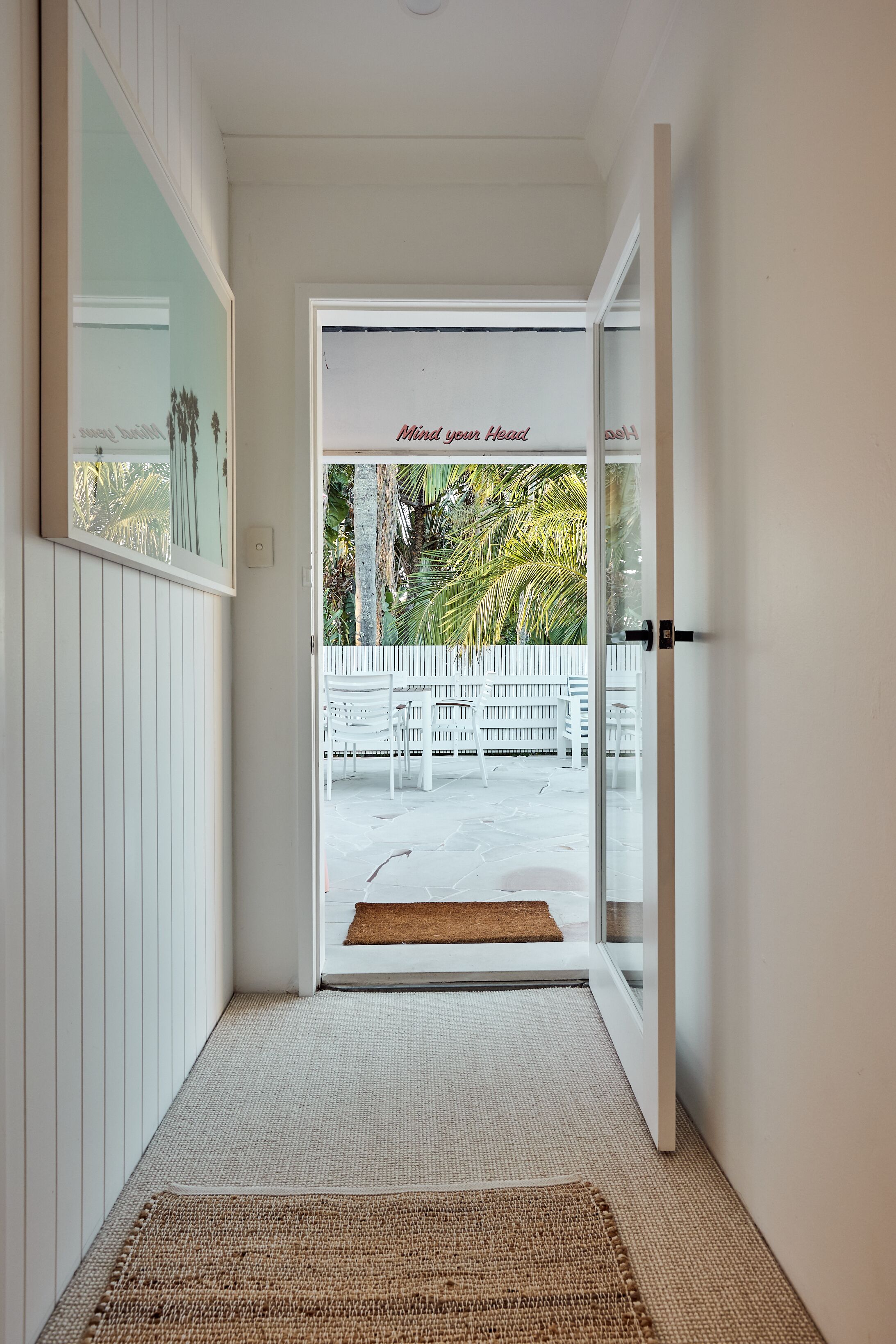 A hallway leading to a glass door with a view of a white fence and palm trees outside.