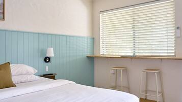 A bedroom with a bed, a nightstand, a lamp, a window with blinds, and two bar stools.