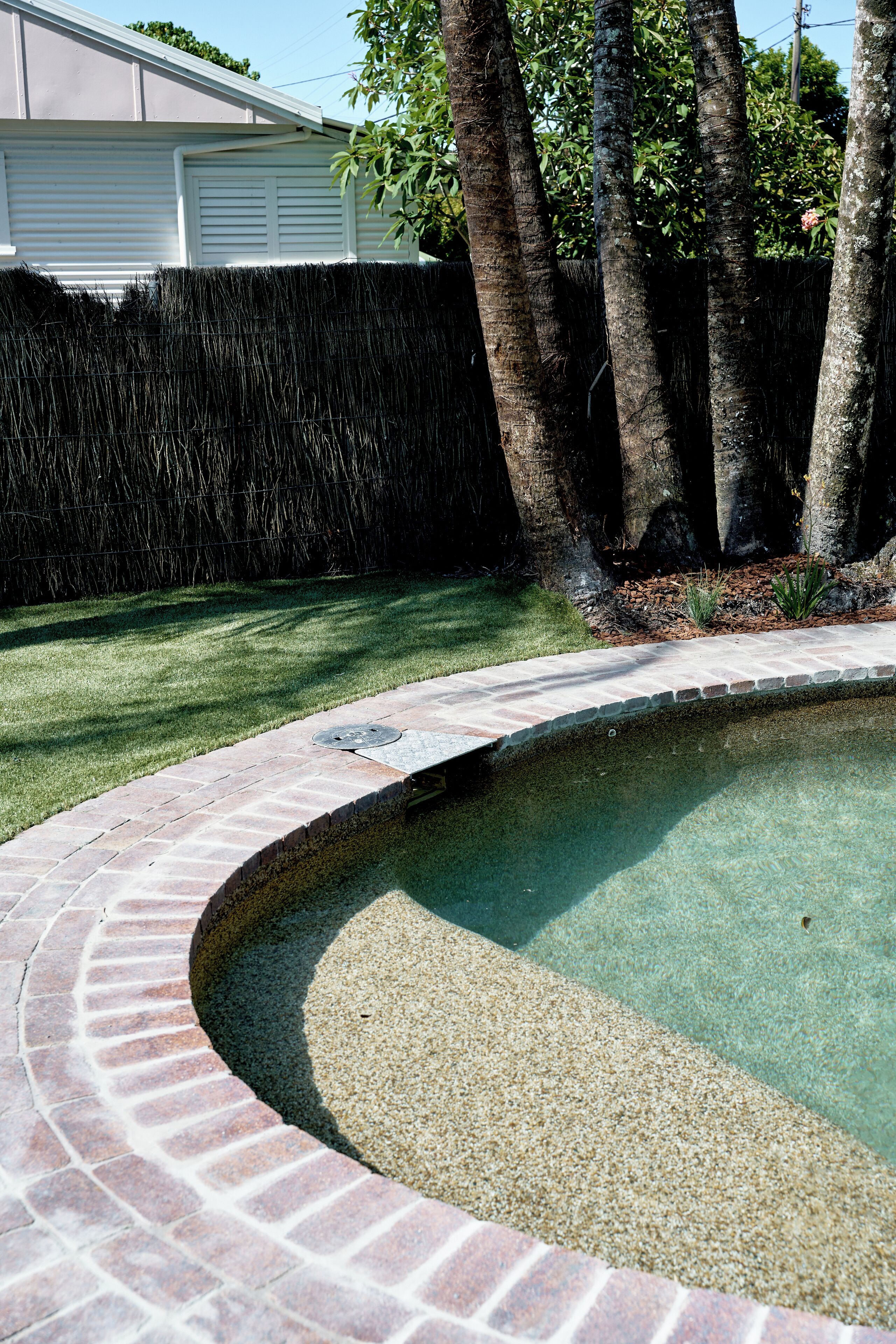Une piscine bordée de briques et une zone herbeuse avec des arbres.