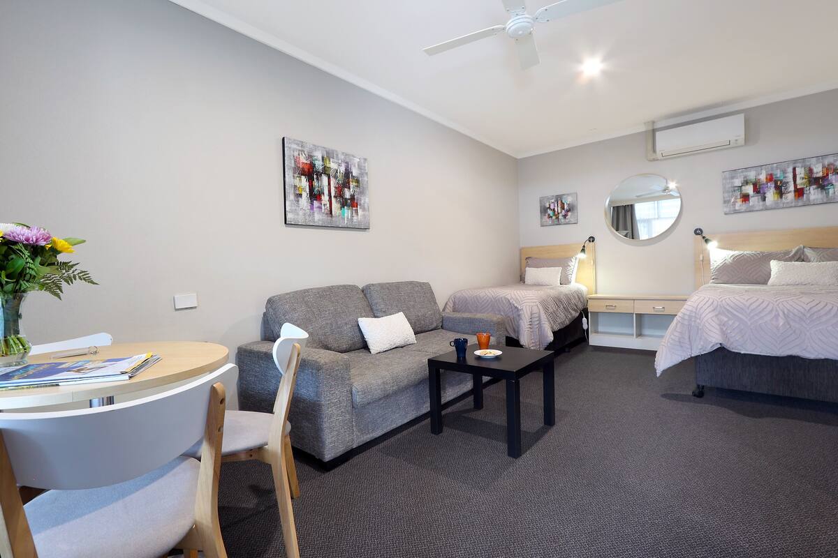 A modern hotel room with a grey sofa, a round dining table, a single bed, and a ceiling fan.