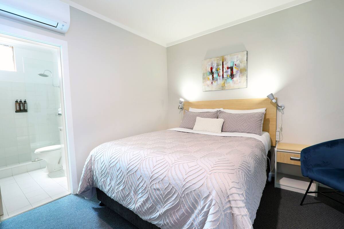 A bedroom with a bed, a bedside table, a chair, a shower, and a toilet.