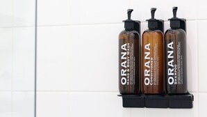 Trois bouteilles de savon pour les mains et le corps de la marque Orana, posées sur une étagère noire devant un mur recouvert de carreaux blancs.