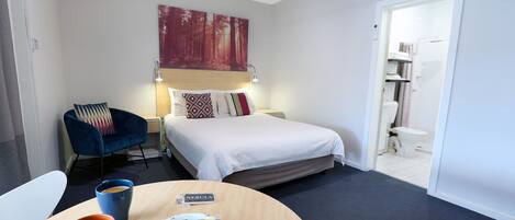 Habitación de hotel con una cama, un escritorio con una taza de café y aperitivos, una silla y un cuadro en la pared.