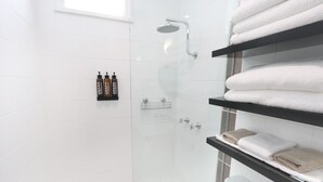 Une salle de bain moderne avec des carreaux blancs, une douche en verre et des étagères où sont rangées les serviettes et les articles de toilette.