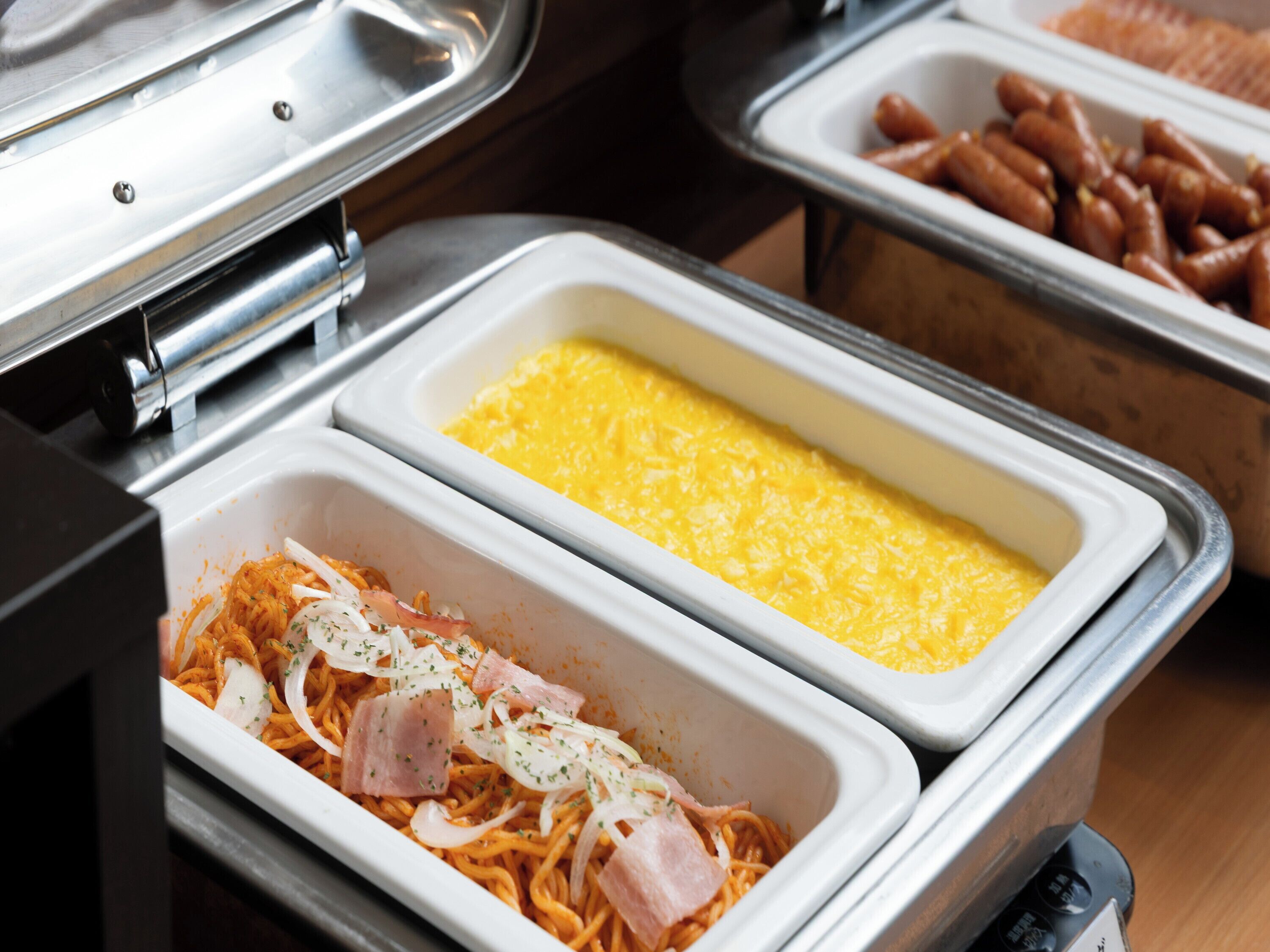 A buffet with various food options including pasta, scrambled eggs, and sausages.