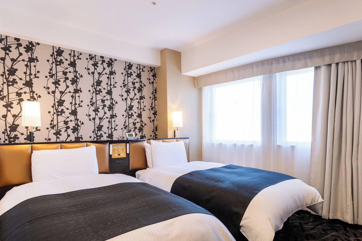 A hotel room with two beds, a floral wall design, a control panel, and a large window with curtains.