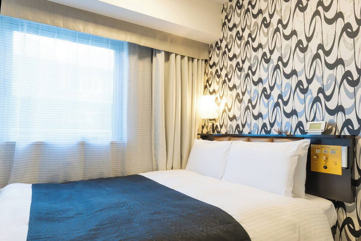 A hotel room with a bed, curtains, a patterned wall, a nightstand, and a lamp.