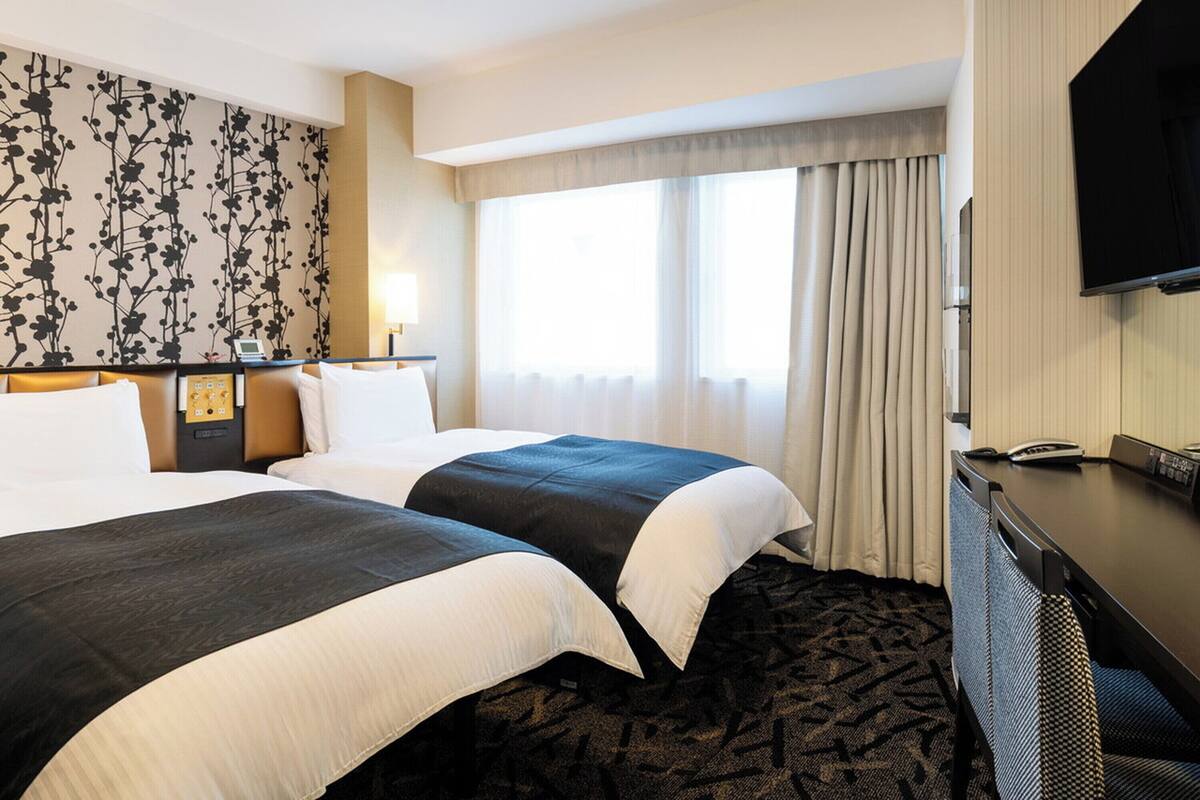 A hotel room with two beds, a desk with a telephone, a flat-screen TV, and a window with curtains.