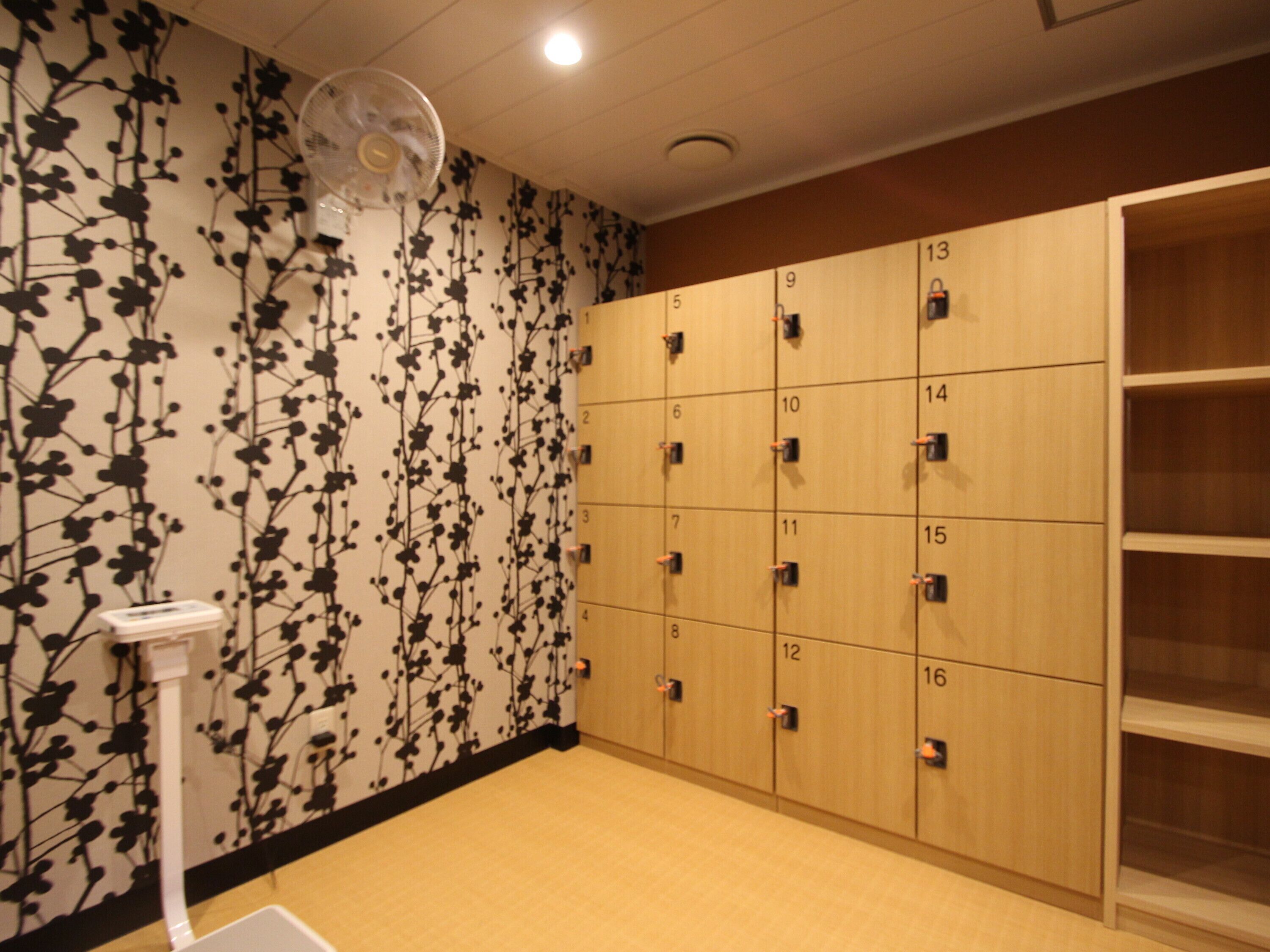 A locker room with numbered lockers, a wall-mounted fan, and a wooden cabinet with shelves.