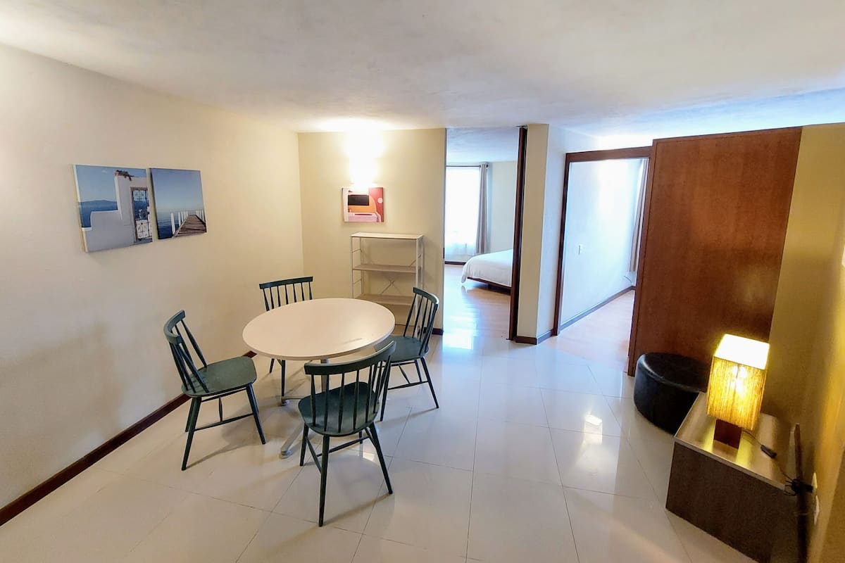 A dining area with a round table and four chairs, a small shelf unit, and a bed in the adjacent room.