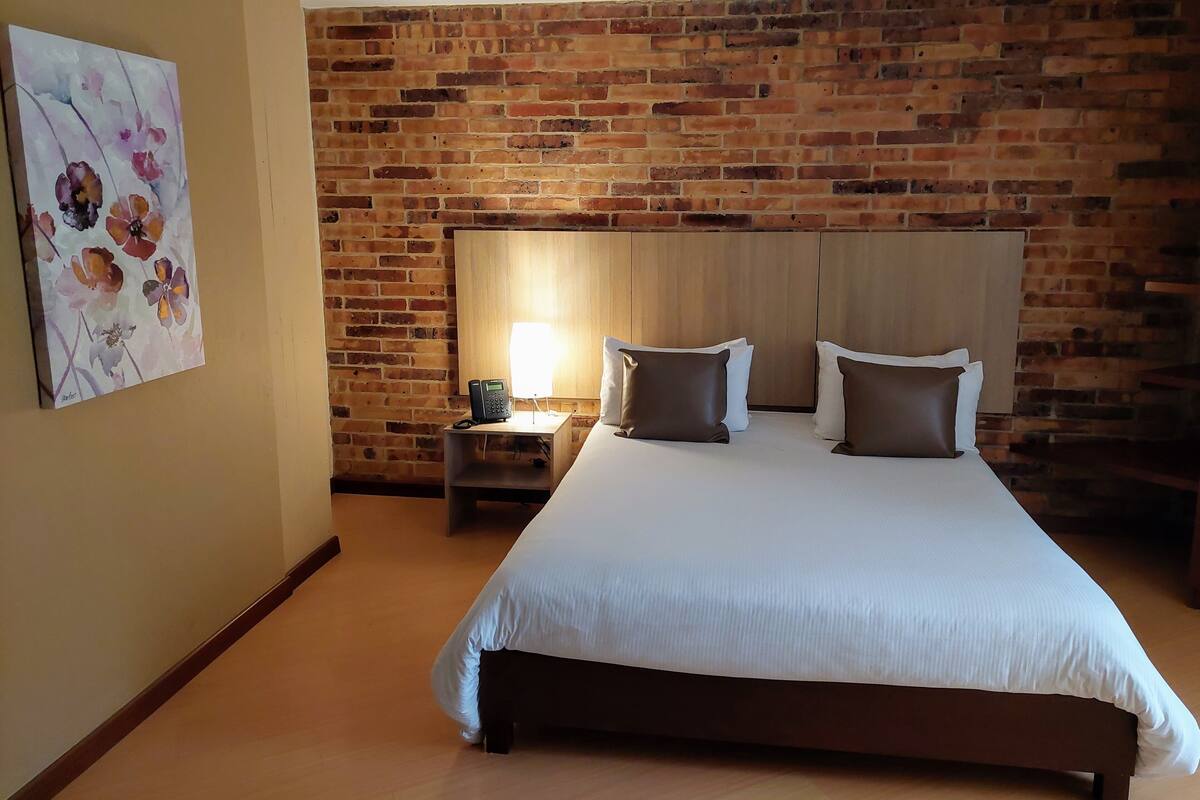 A hotel room with a brick wall, a bed with white bedding, two pillows, a bedside table with a lamp, and a phone.