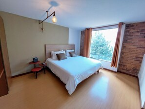 A neatly arranged bedroom with a large bed, a small bedside table, a lamp, and a window with curtains. - Viaggio Urbano (Bogotá)