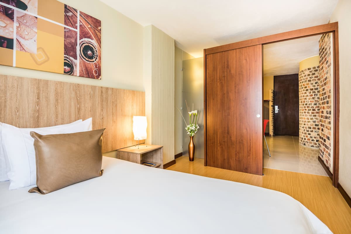 A hotel room with a large bed, a nightstand with a lamp, a vase with flowers, and a wooden wardrobe.
