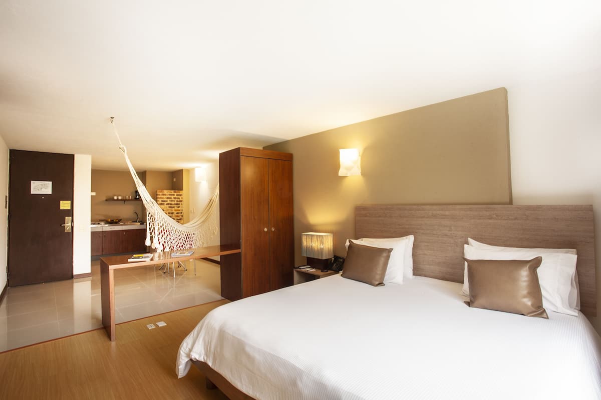A hotel room with a large bed, a wooden wardrobe, a desk with a chair, a hammock, and a small kitchen area.