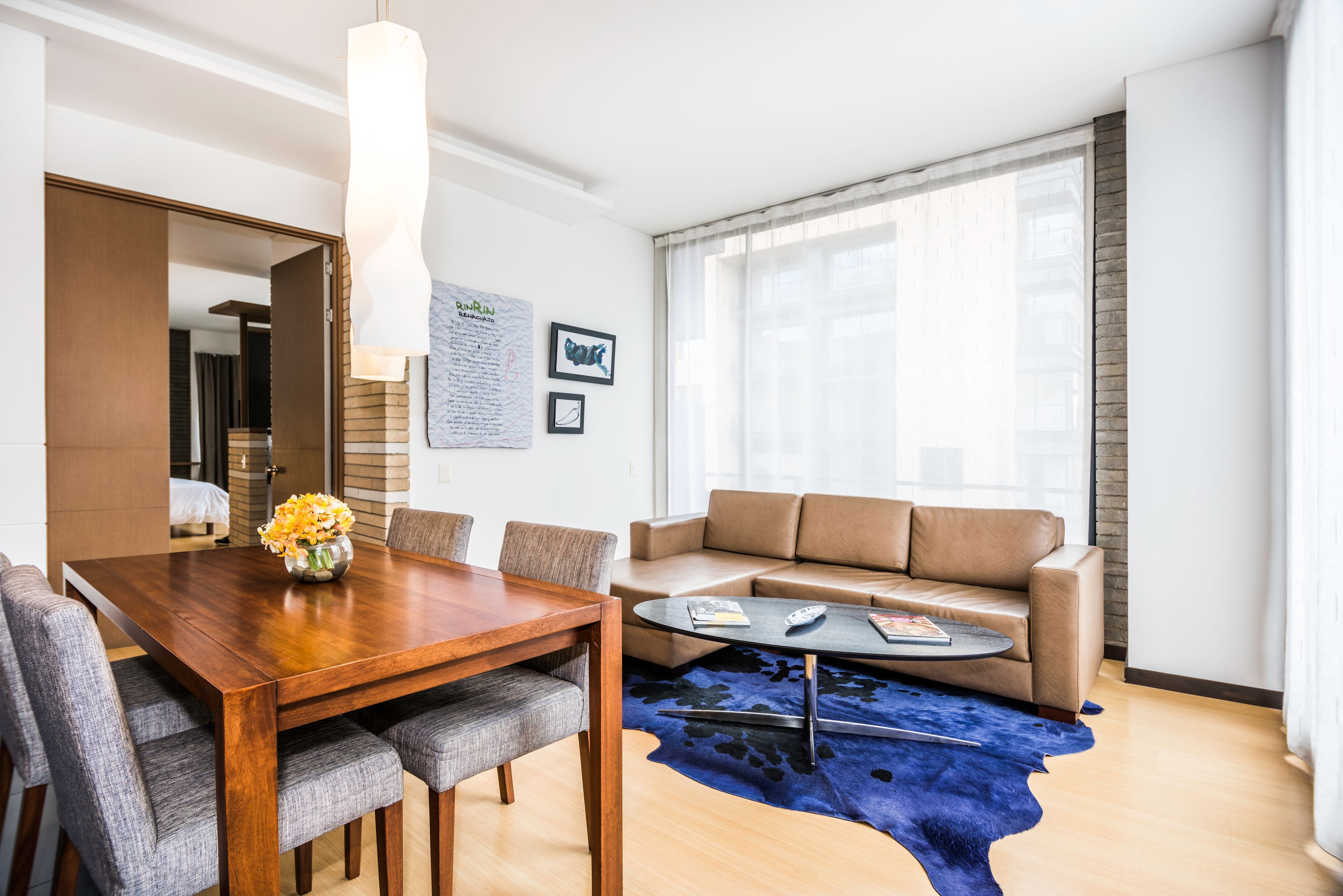 Een moderne woonkamer met een houten eettafel, grijs gestoffeerde stoelen, een bruine leren bank, een ronde salontafel en een blauw vloerkleed.