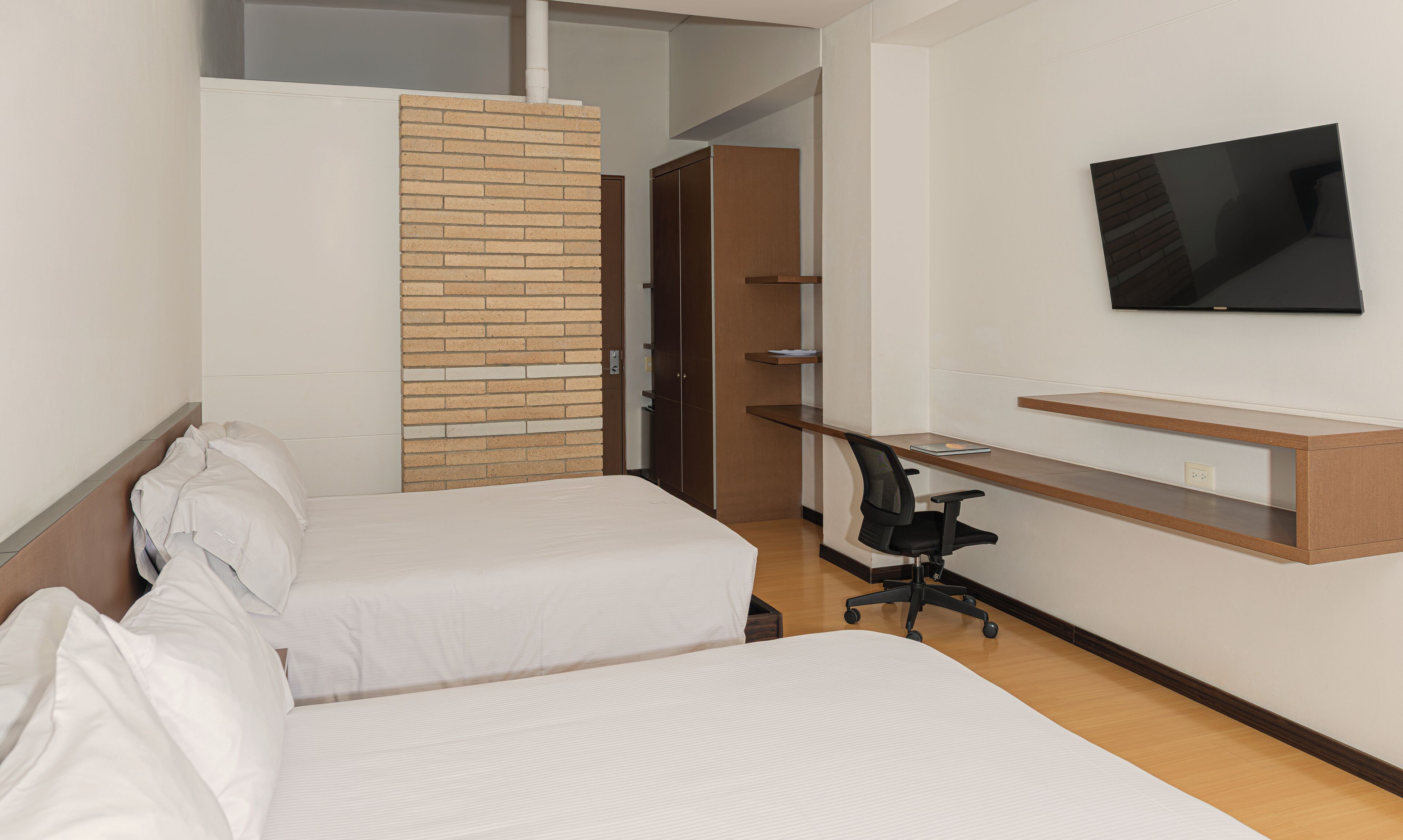 A hotel room with two beds, a desk, a chair, a TV, and a brick accent wall.