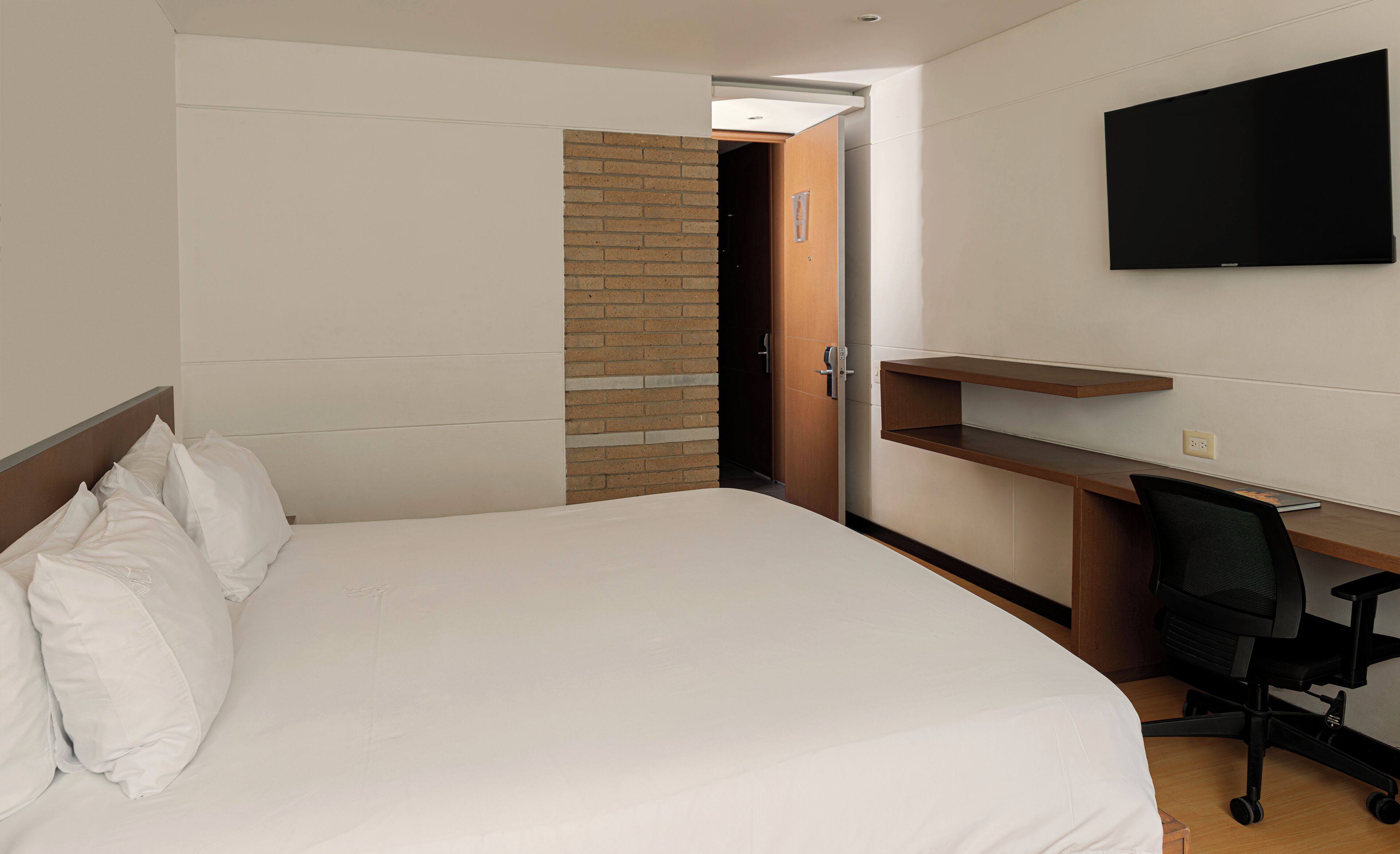 A hotel room with a bed, a desk, a chair, a TV, and a brick wall.
