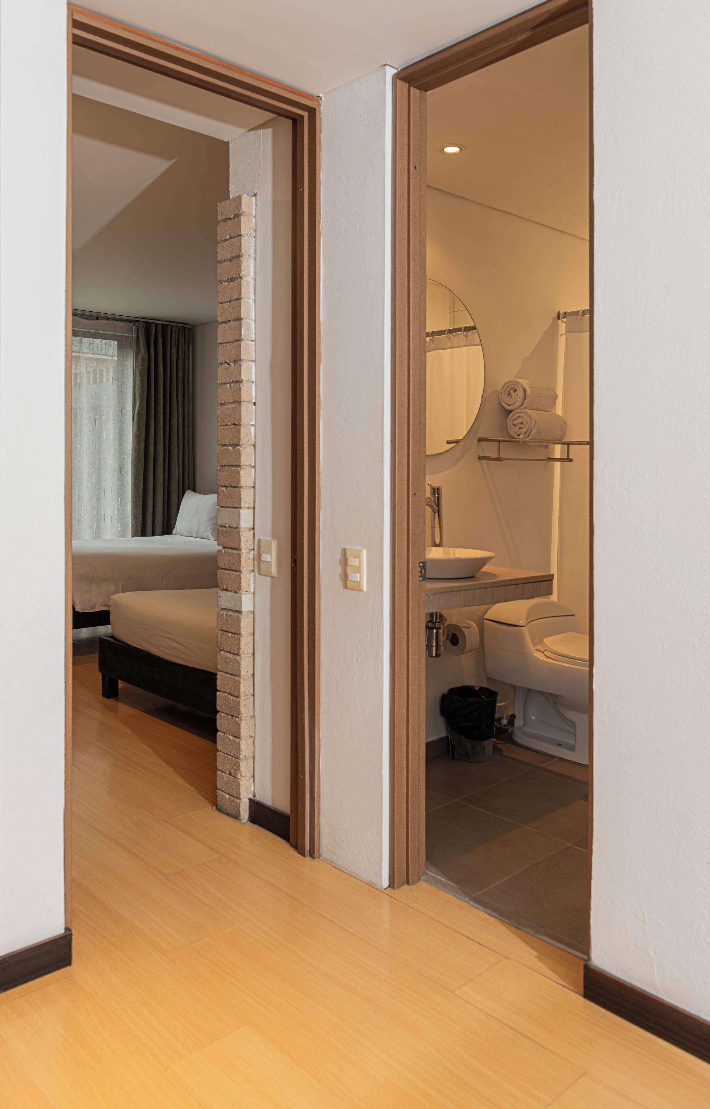 A hotel room with a bed, a bathroom with a sink and toilet, and a hallway with wooden flooring.